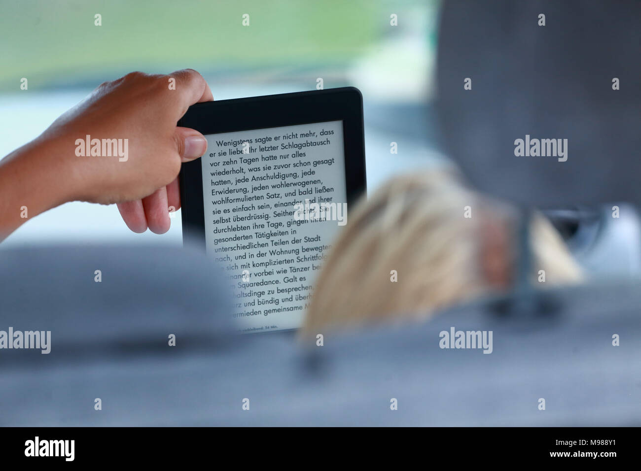 Rubio womand desde detrás sentado en el asiento del pasajero en un coche leyendo un libro alemán sobre un e-reader Foto de stock