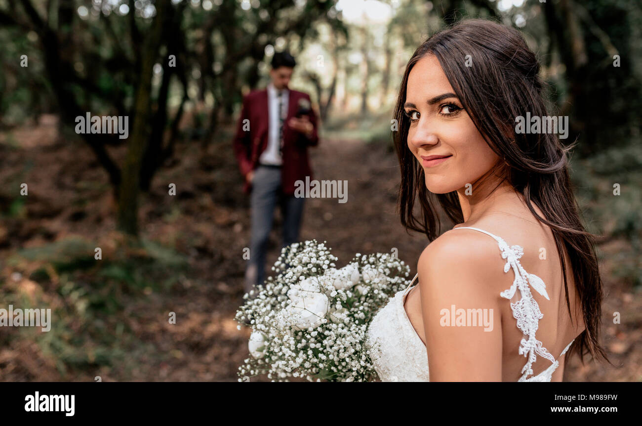 Retrato de novia sonriente sosteniendo el ramo de flores en el bosque con el novio en segundo plano. Foto de stock