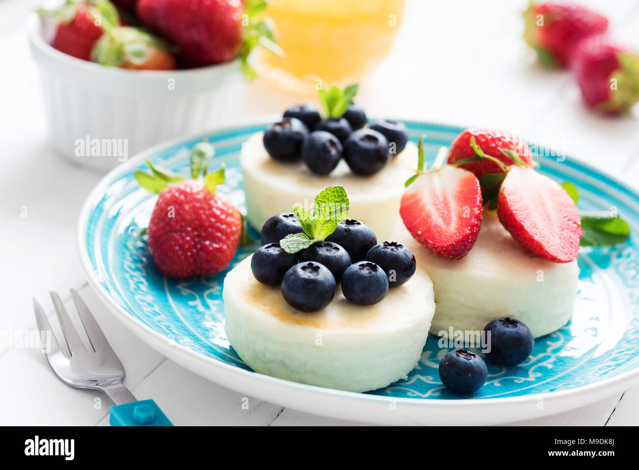 Mini cheesecake con fresas frescas y arándanos sobre placa azul, acercamiento. Tortitas, queso cottage Syrniki Foto de stock