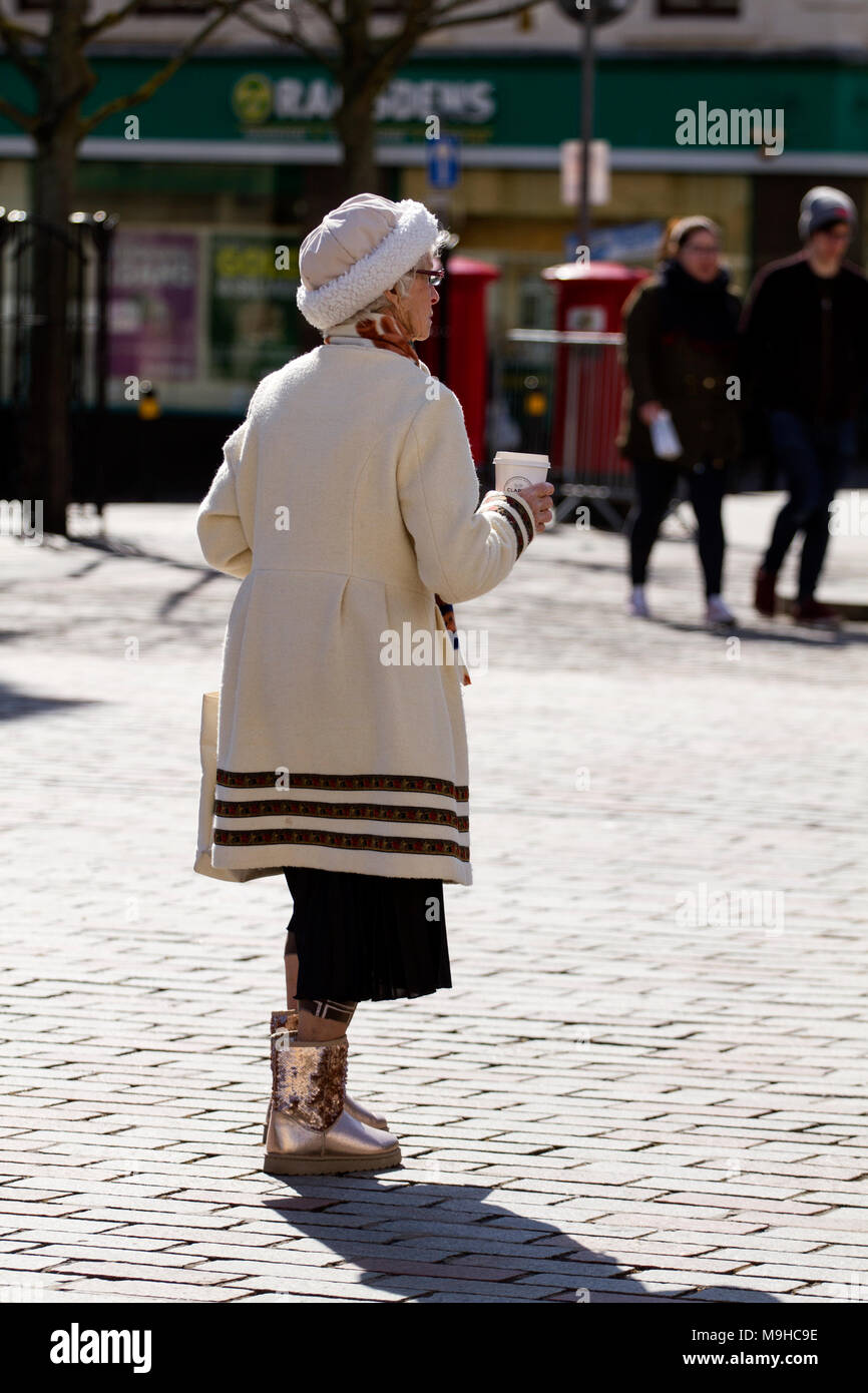 Una moda mujer anciana sosteniendo una taza de café de Clark's Bakery y  luciendo brillantes Ugg botas caminando en el centro de la ciudad escocesa  de Dundee, Reino Unido Fotografía de stock -