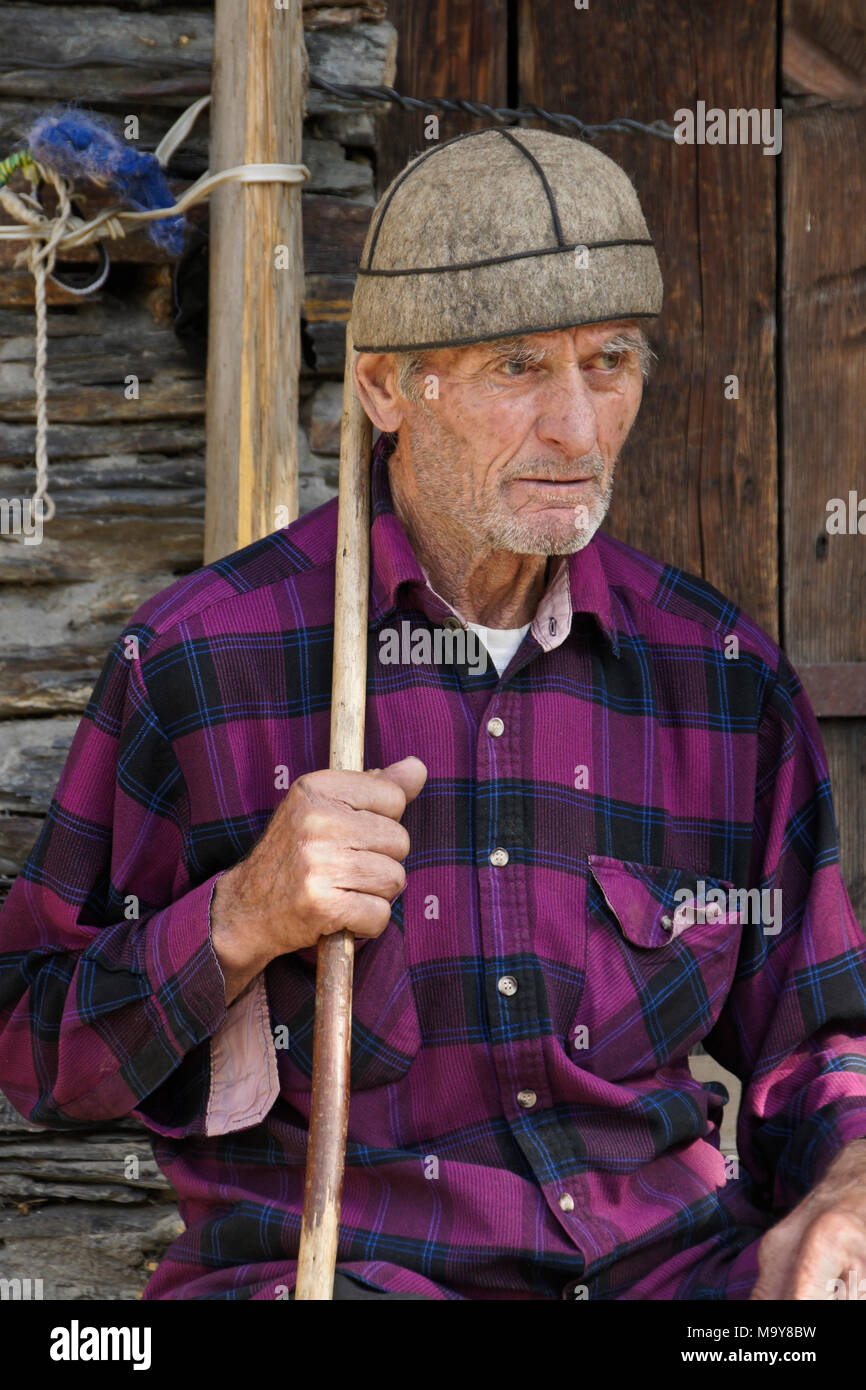 Un anciano sentada fuera un cobertizo de piedra, llevaba una camisa de cuadros escoceses y tapón de lana, bastón por su lado, Ushguli, Superior Svaneti, Georgia Foto de stock