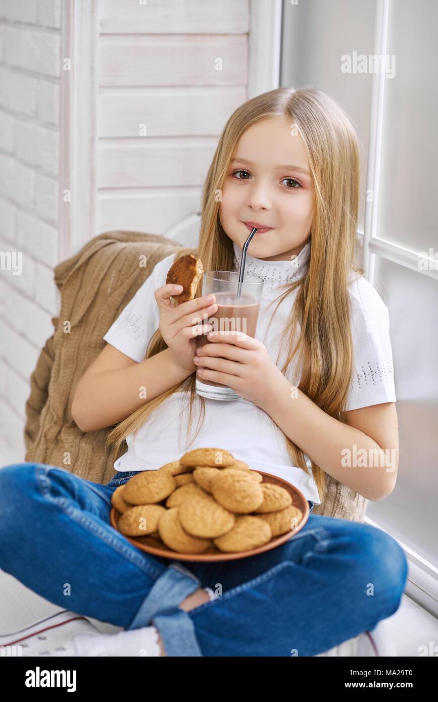 Nina Esta Sentado En Una Silla Vistiendo Pantalones De Mezclilla Y Camiseta Blanca Nino Bebe Leche Con Chocolate De Vidrio Transparente Y Tiene Gran Plato Lleno De Deliciosas Galletas Dulces Sobre Sus