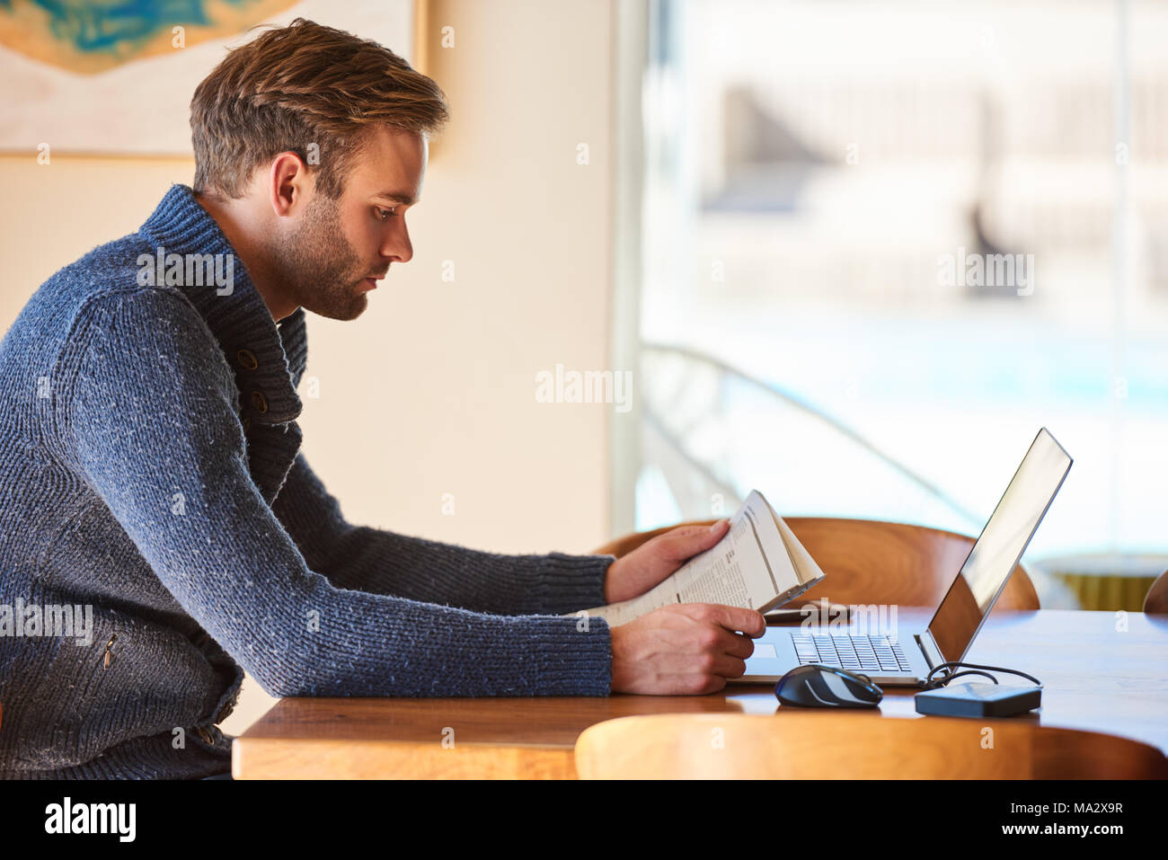 Hombre blanco sentado en la mesa del comedor temprano en la mañana ocupado leyendo su diario para ponerse al día sobre asuntos actuales, con su taza de café Foto de stock