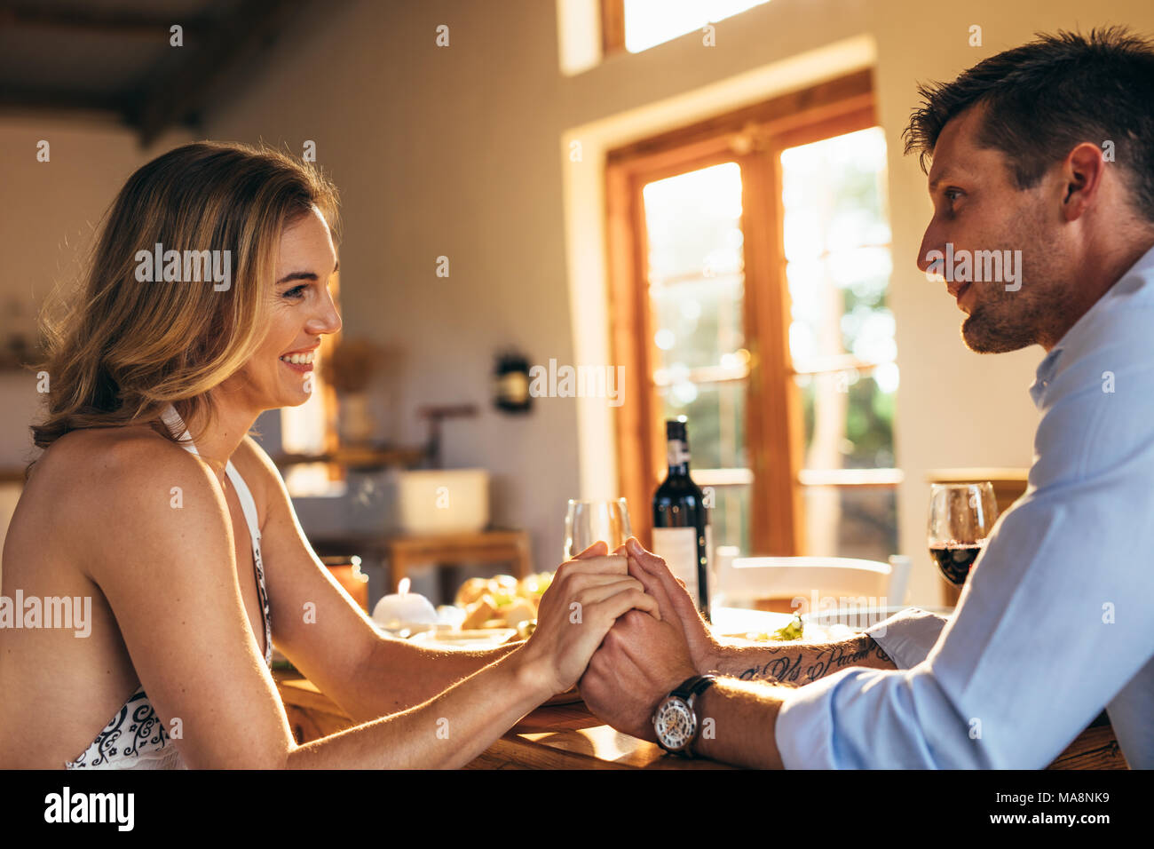 Amorosa pareja joven sentado en la mesa de comedor sosteniendo las manos de cada uno. Un hombre y una mujer con una cena romántica en casa. Foto de stock
