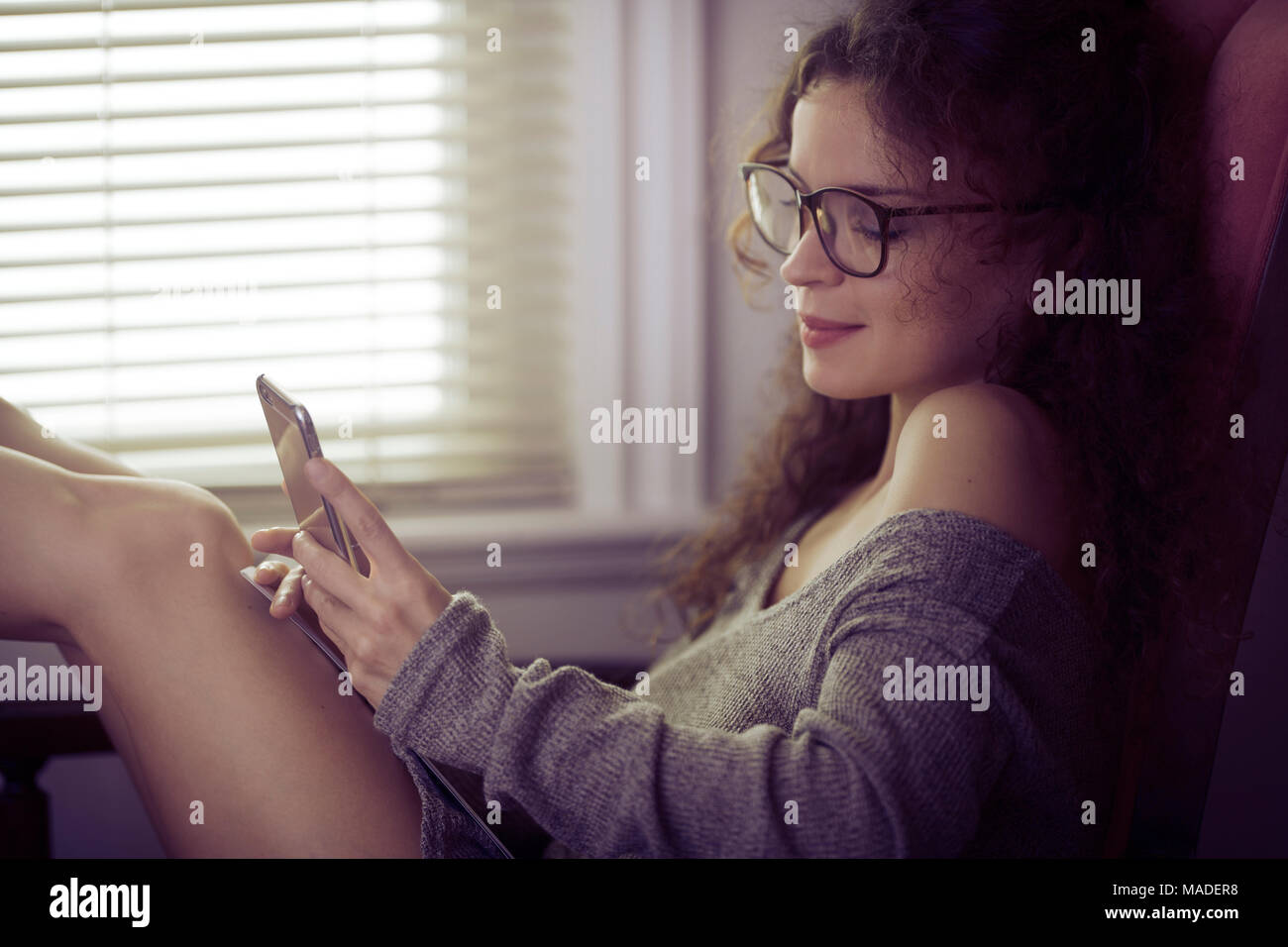 Retrato de una joven mujer sonriente en gafas para leer sentado con un iPhone en sus manos en una silla junto a la ventana, los mensajes de texto, mensajería concepto Foto de stock