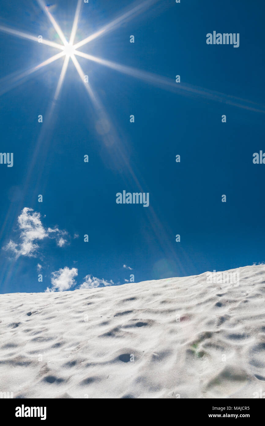 Colina cubierta con nieve en las montañas con sun bengalas sobre el cielo azul, con algunas pequeñas nubes Foto de stock