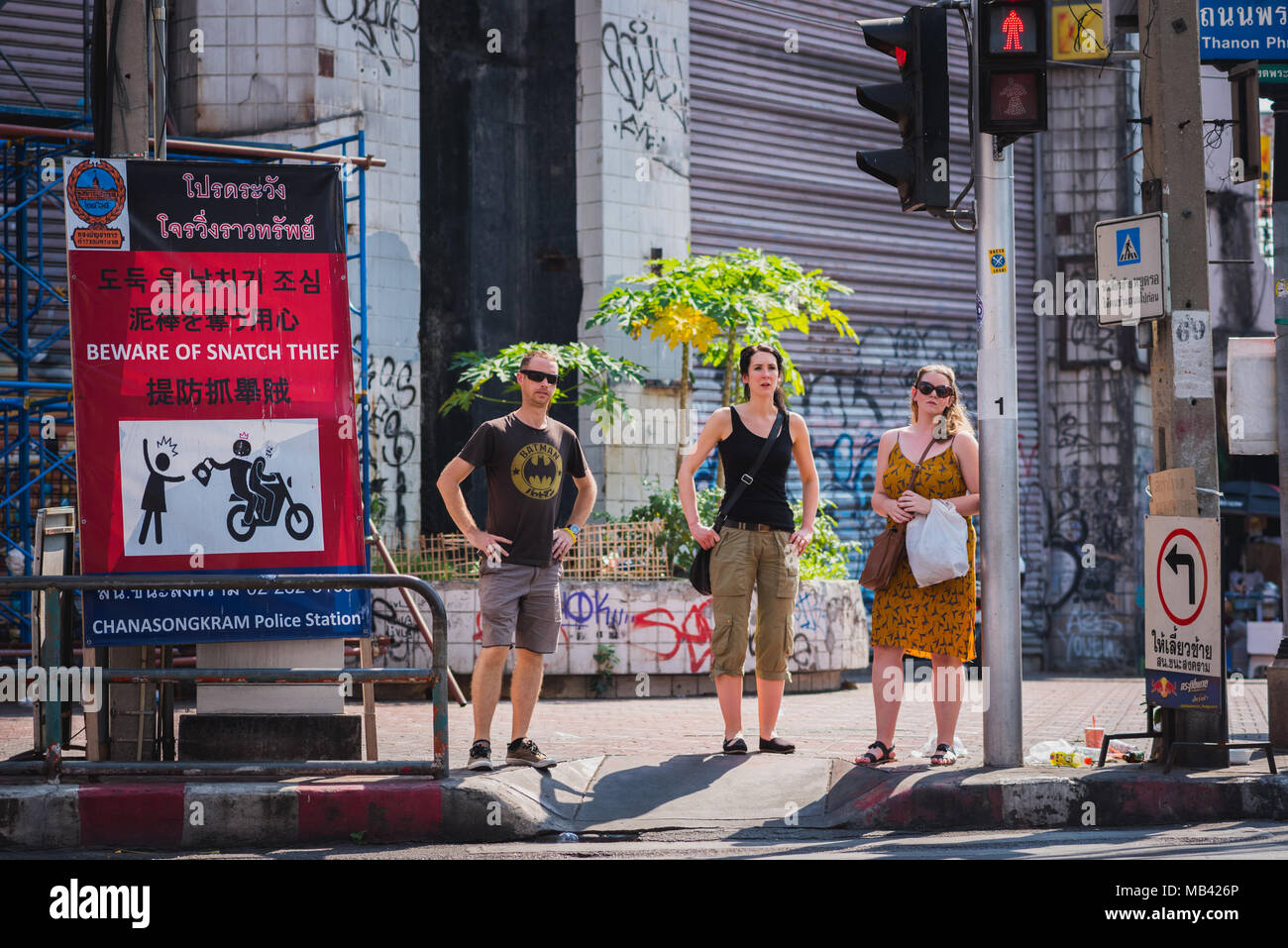 Tres mochileros situarse en el cruce de peatones tras las vacaciones de verano ropa sobre ellos. Hay una señal que advierte acerca de arrebatar los robos en Bangkok Foto de stock