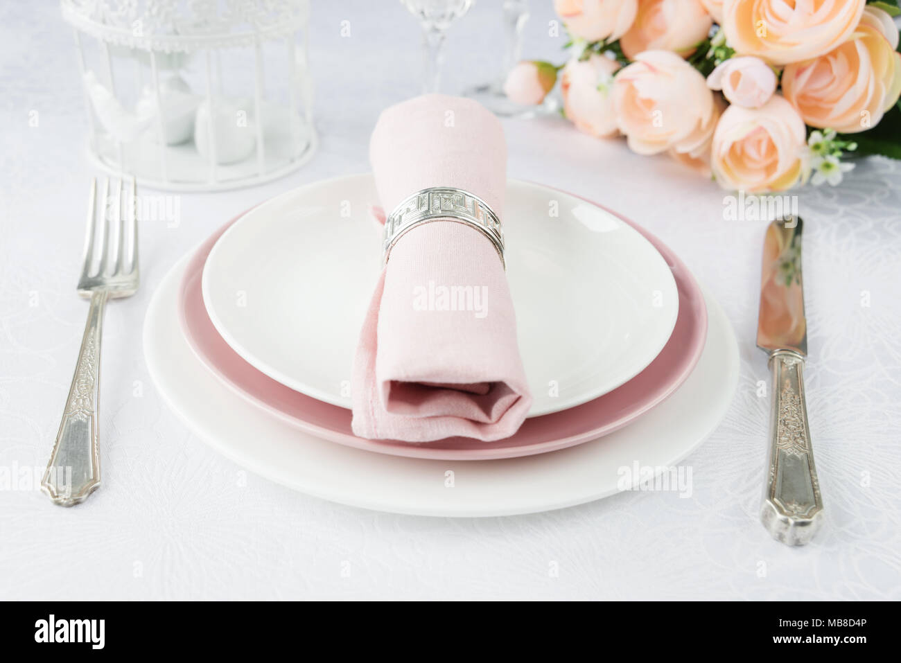Tabla bellamente decorada con blanco y rosa, platos, vasos, cubiertos, servilleta rosa y flores sobre manteles de lujo Foto de stock
