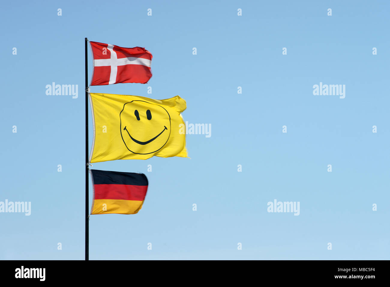 Ondeando banderas alemanas y danesas junto con un smiley bandera en un asta de bandera Foto de stock