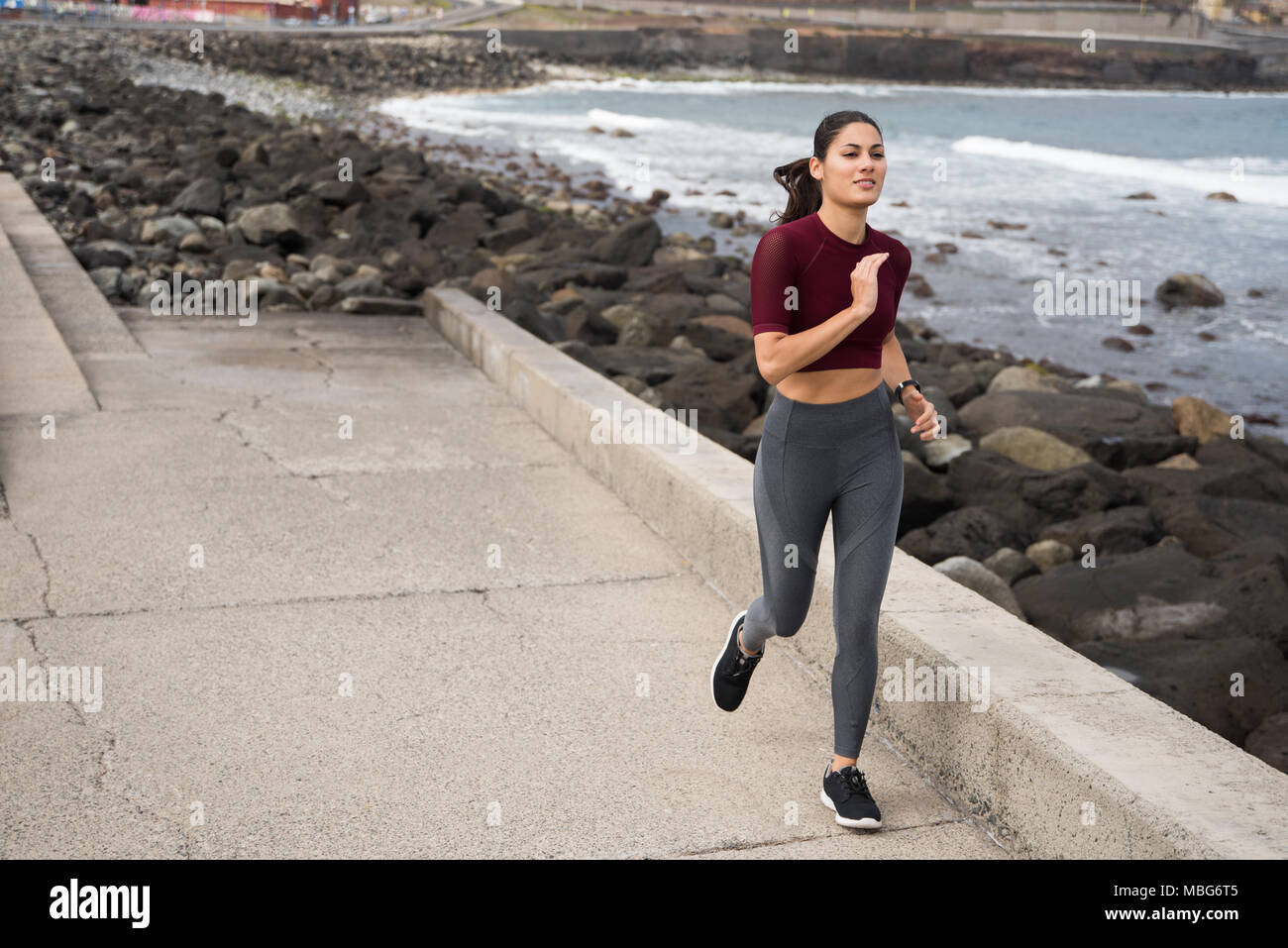 Hermosa morenita corriendo por la orilla del mar en ropa deportiva con su cabello amarrado atrás Foto de stock