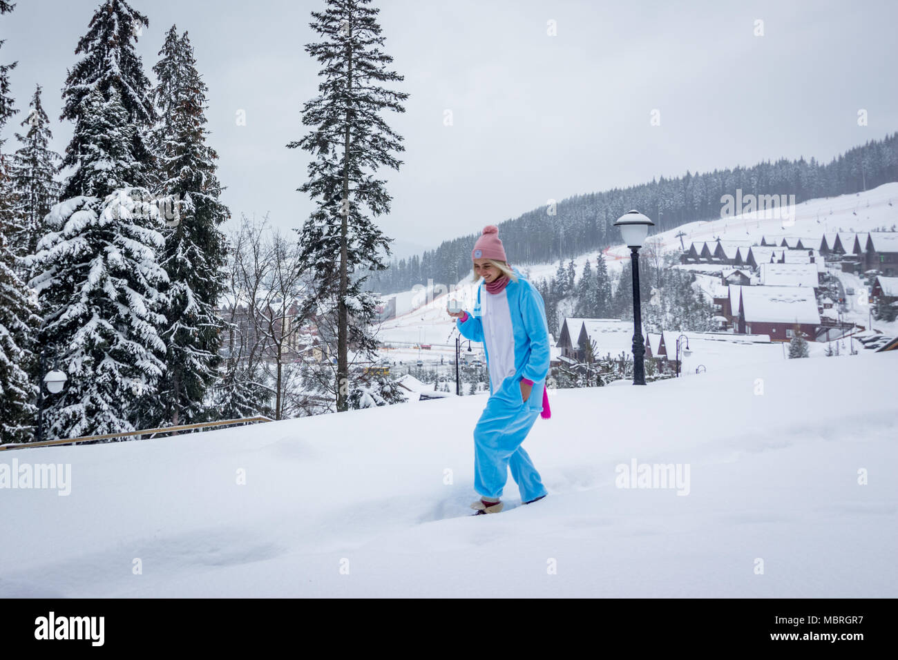 Chica en azul, rosa unicorn pijama kigurumi piscina en frente de las casas de madera sobre el informe de esquí en las montañas de nieve bebiendo café Foto de stock