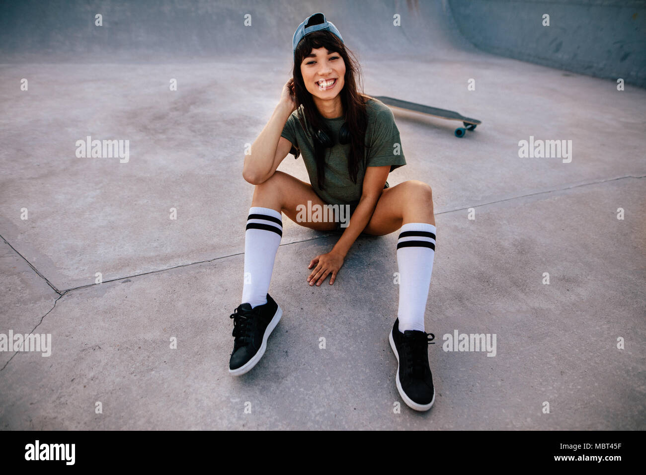 Retrato de cool joven skateboarder sentado en el parque de skate mirando a la cámara y sonriendo. Hipster mujer relajarse al aire libre en el parque de skate. Foto de stock