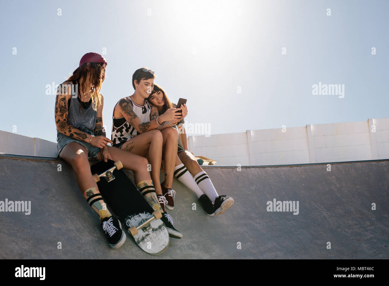 Grupo de amigas sentado en la rampa en el parque de skate y mirando al  teléfono móvil. Tres mujeres skaters utilizando smart phone junto al parque  de skate Fotografía de stock -