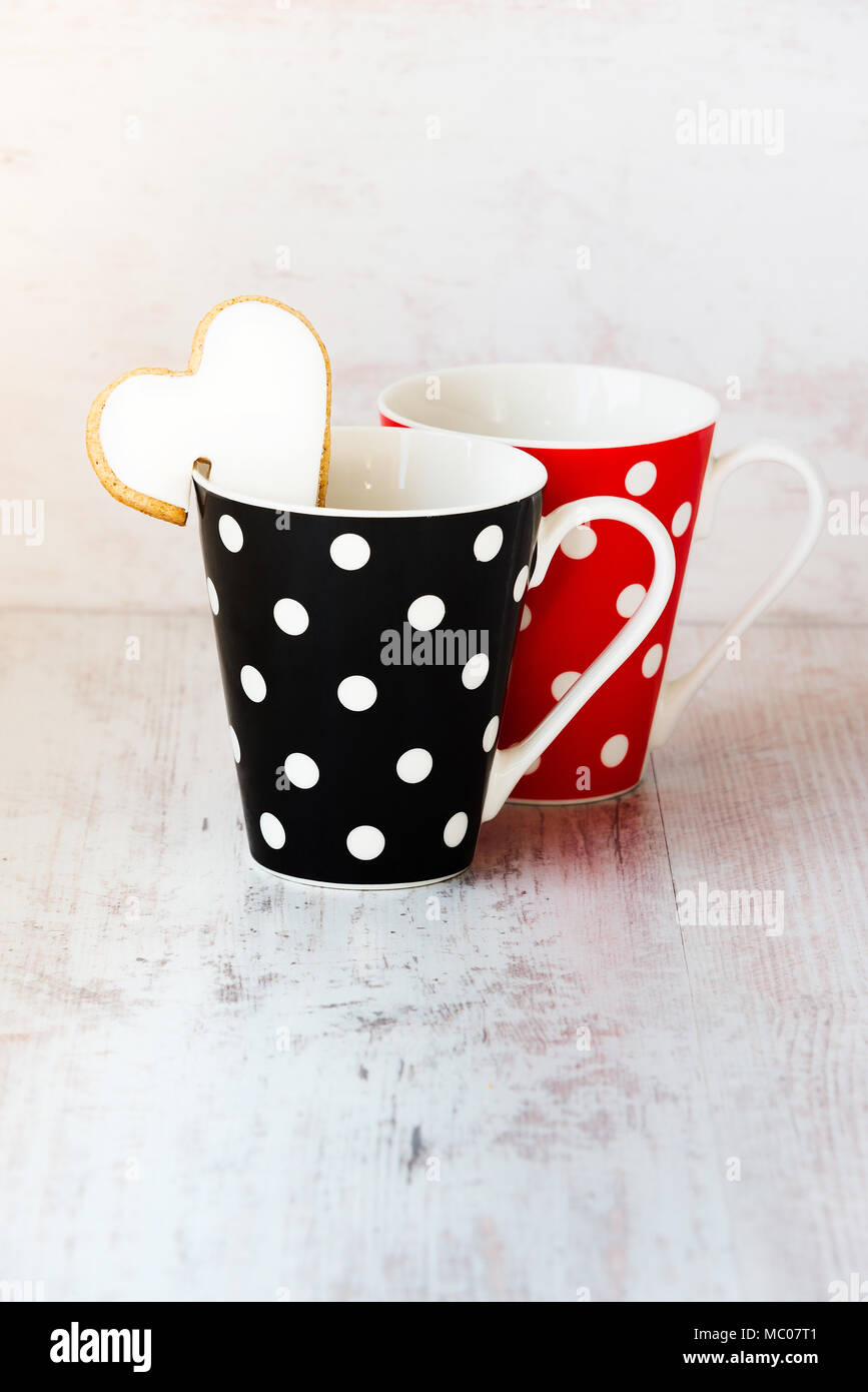 Un par de negro y rojo puntos polka tazas de café con galletas caseras en forma de corazón en el borde sobre la madera blanca de fondo. Foto de stock