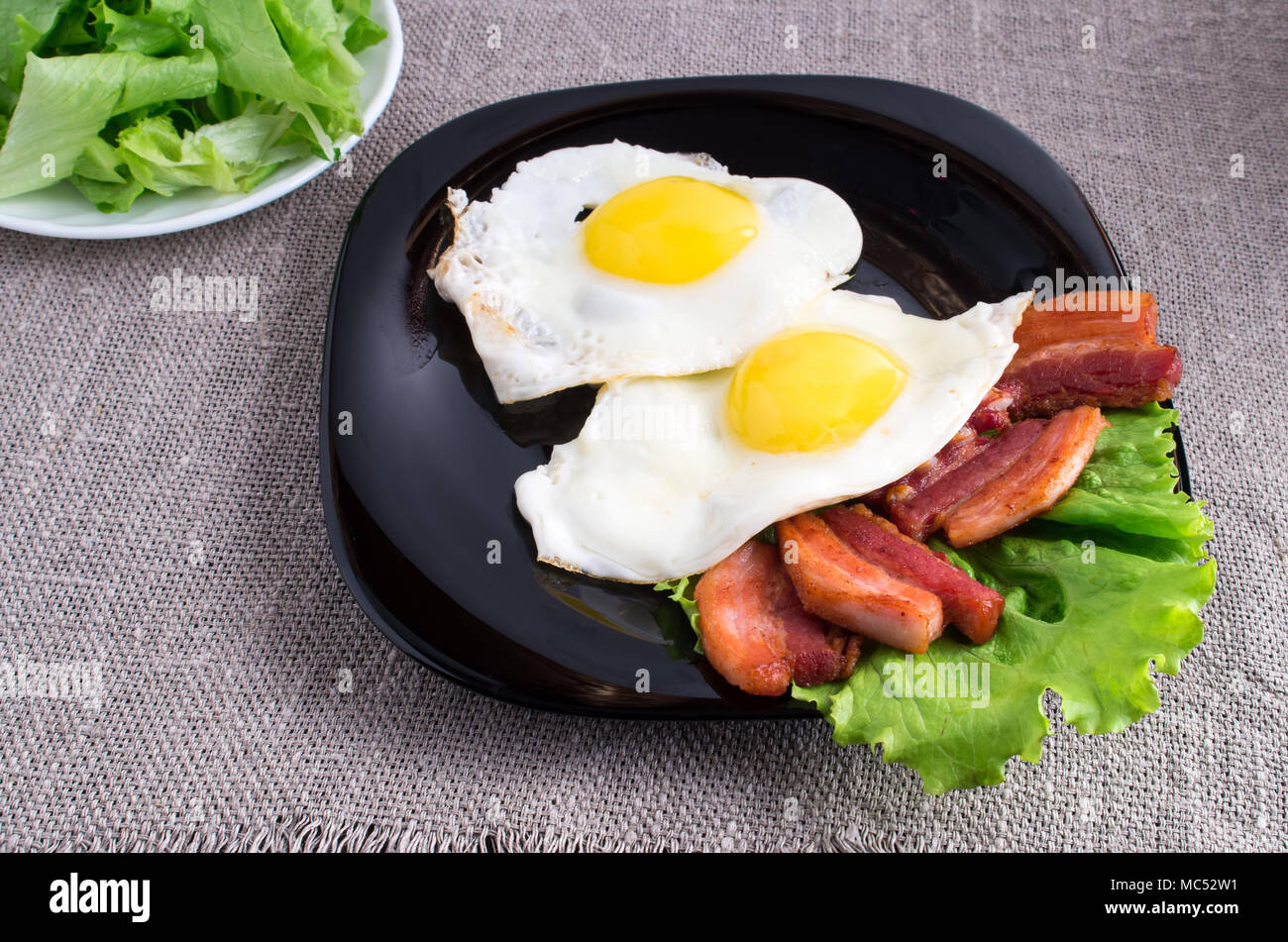 Placa negra con huevos fritos, tocino y lechuga closeup sobre un mantel gris Foto de stock