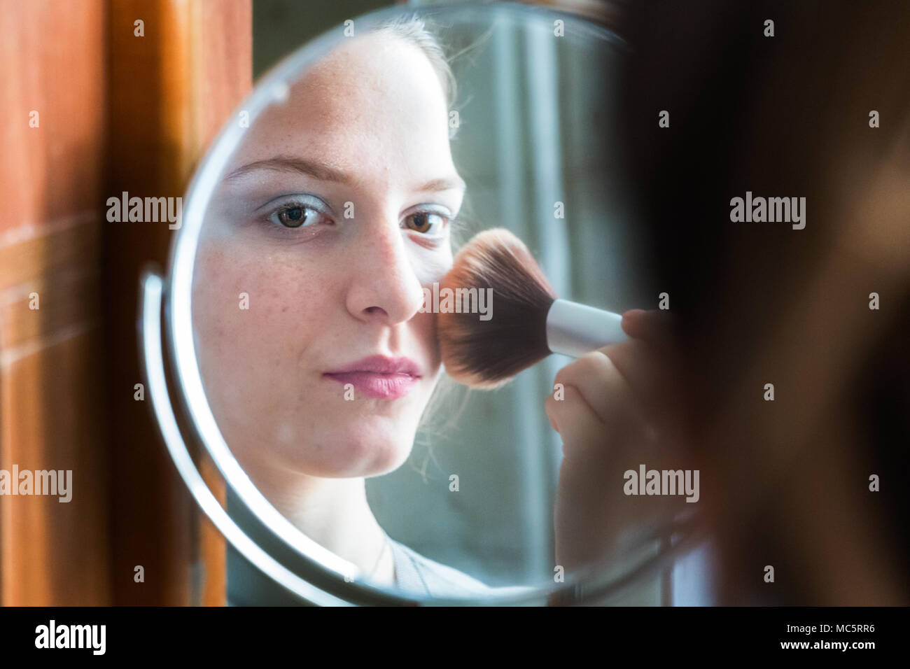 Joven aplicando el polvo con un cepillo, mirando su reflejo en un espejo redondo en casa Foto de stock