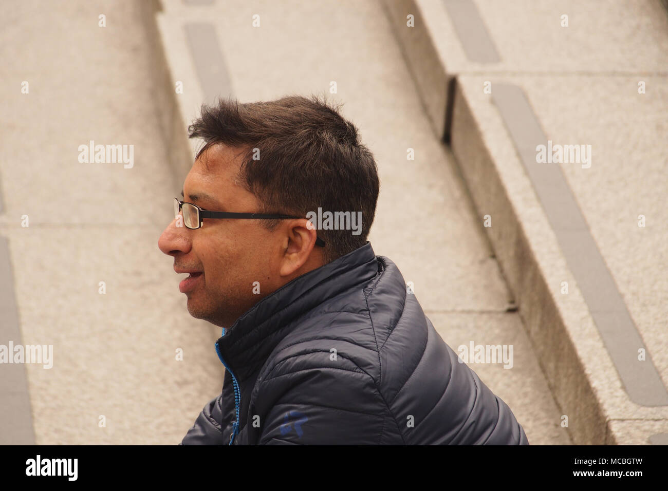 Un hombre joven sentado en los escalones de piedra en Trafalgar, Londres viendo el mundo pasar vistiendo una capa gruesa y gafas Foto de stock