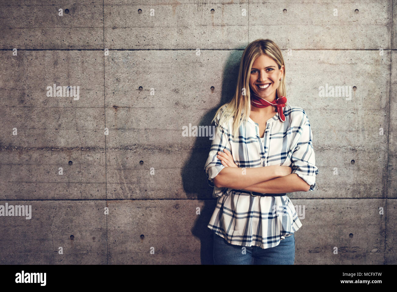 Hermosa joven sonriente exitoso emprendedor femenino de pie delante de un muro de hormigón con manos cruzadas y mirando a la cámara. Foto de stock