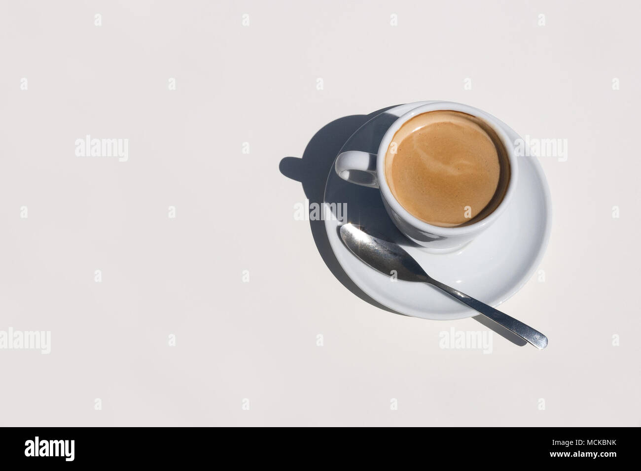 Una deliciosa taza de café caliente en una taza y plato blanco con cuchara sentado en una mesa blanca vista aérea con espacio a la izquierda. Foto de stock