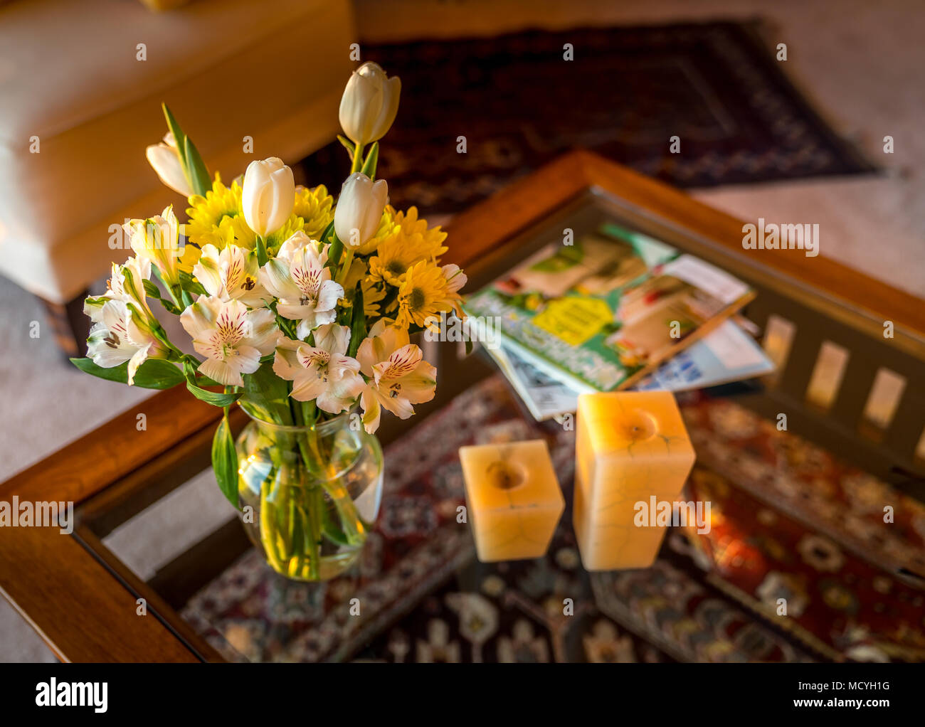 Decoración interior en un ajuste de color cálido; flash las flores cortadas en un frasco de vidrio, la luz de las velas y revistas sobre una mesa de café superior de cristal moderno. El wo Foto de stock