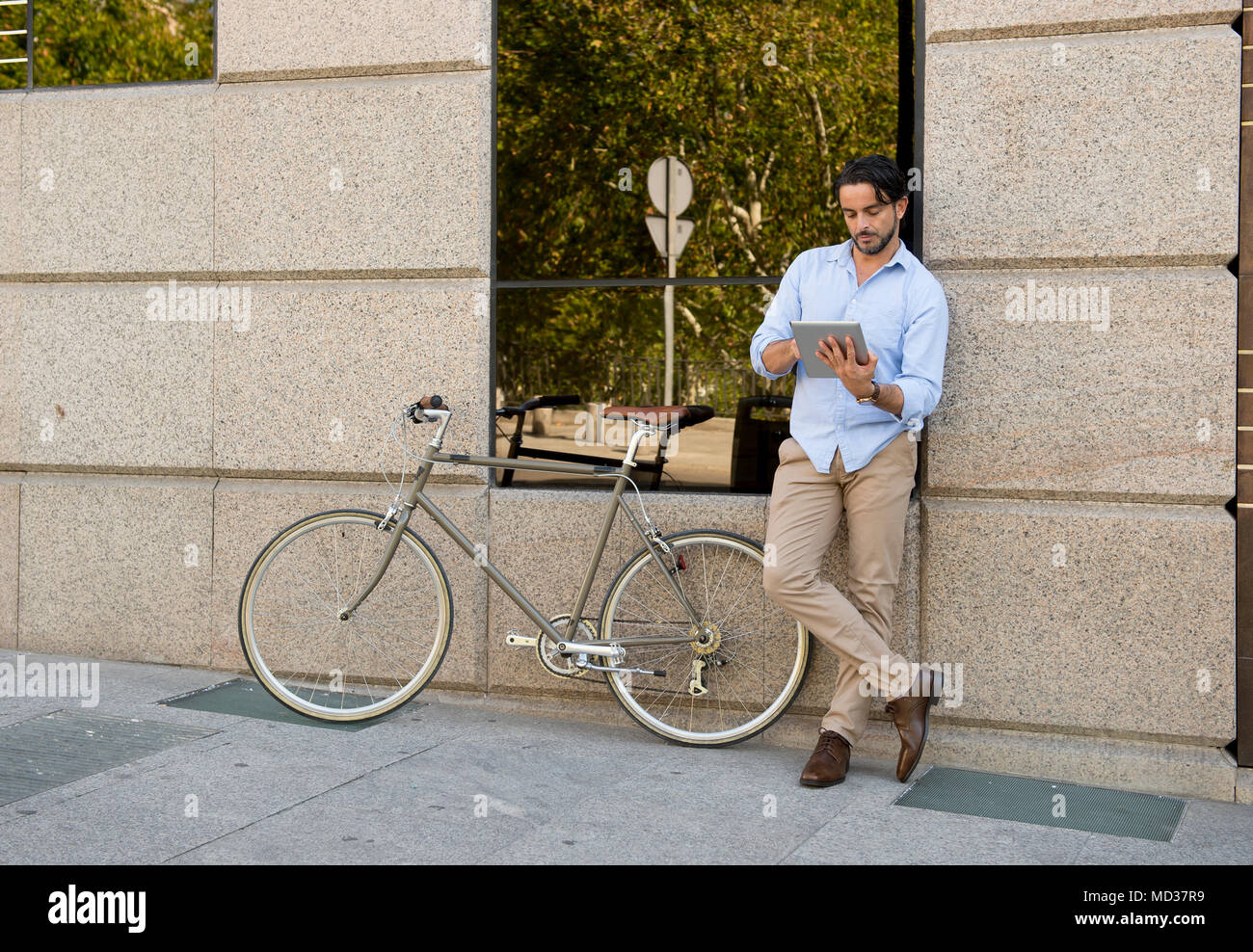 Feliz joven hombre hispano atractiva en ropa de moda casual sonriendo alegre utilizando internet con tablet pad digital en vintage cool retro en bicicleta Fotografía de stock -