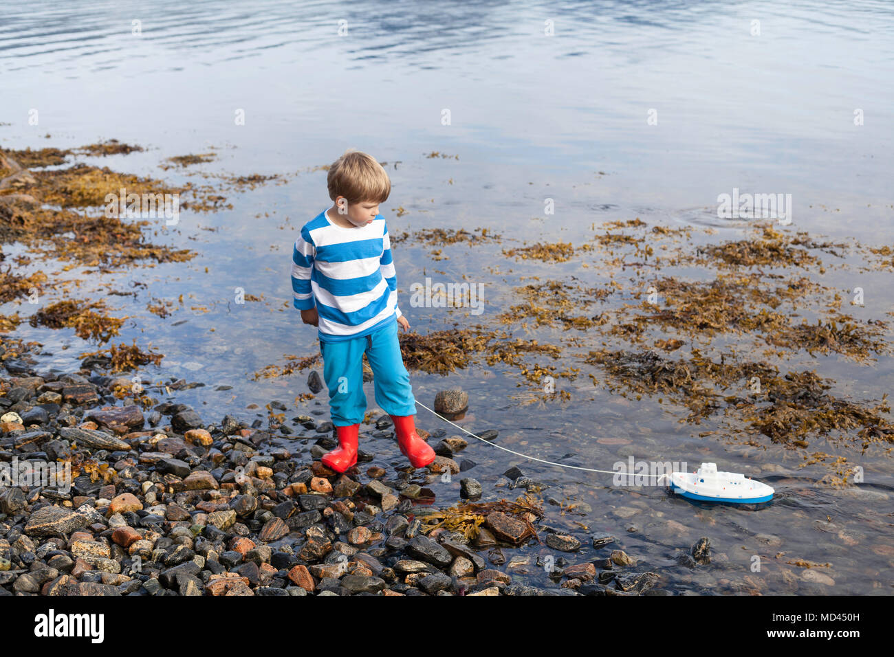 Fiordo Boy en el borde del agua jugando con barco de juguete, Aure, Møre og Romsdal, Noruega Foto de stock
