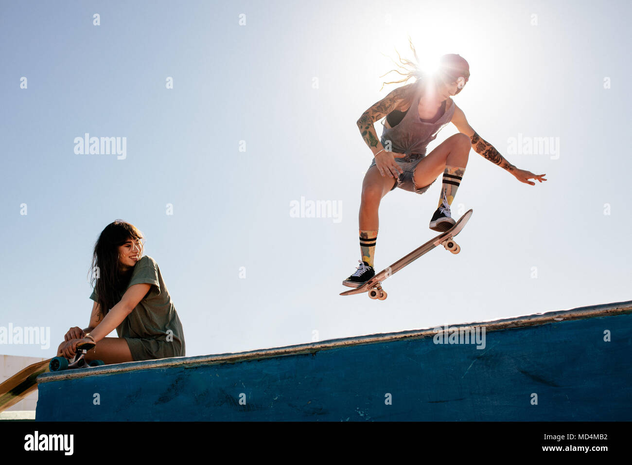 Patinador femenina cabalga sobre monopatín con una amiga sentada en la rampa. Hembra de skate en el skate park con un amigo viendo la rutina. Foto de stock