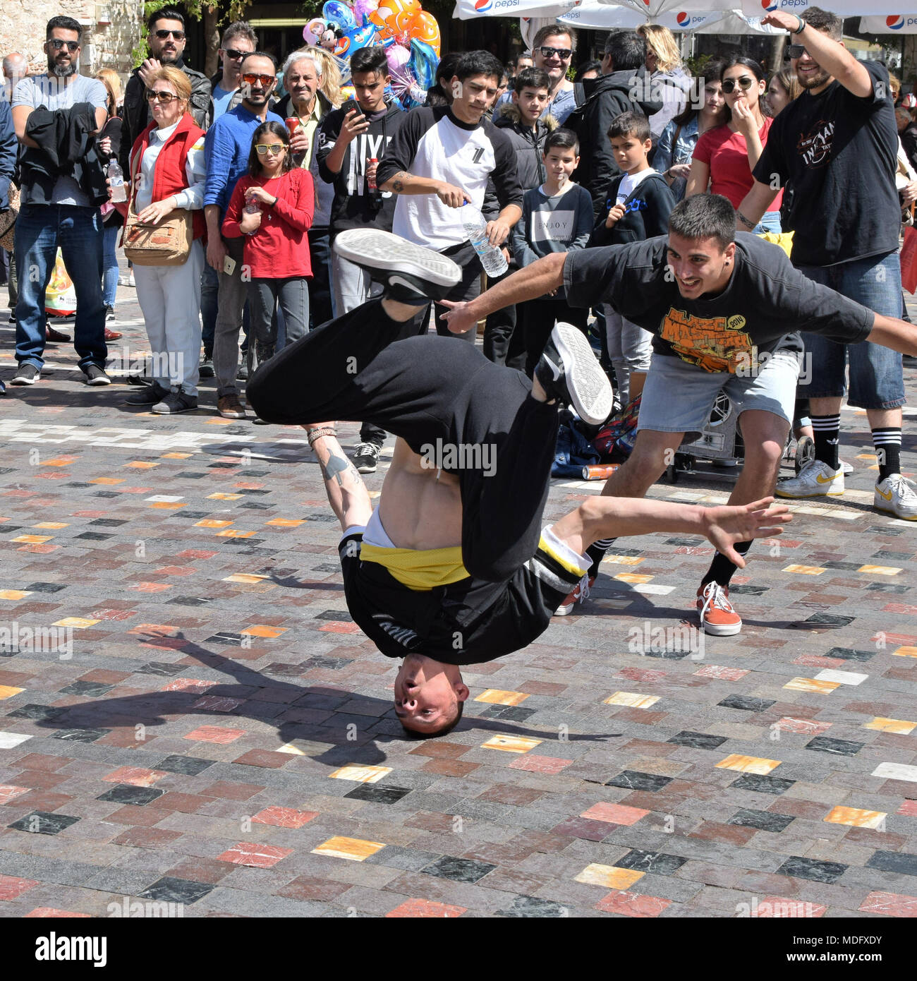 Atenas, Grecia - Abril 1, 2018: Amigos vitoreando en breakdancer como él mueve en headspin bustos una plaza pública con multitud de personas. Juventud street dance. Foto de stock