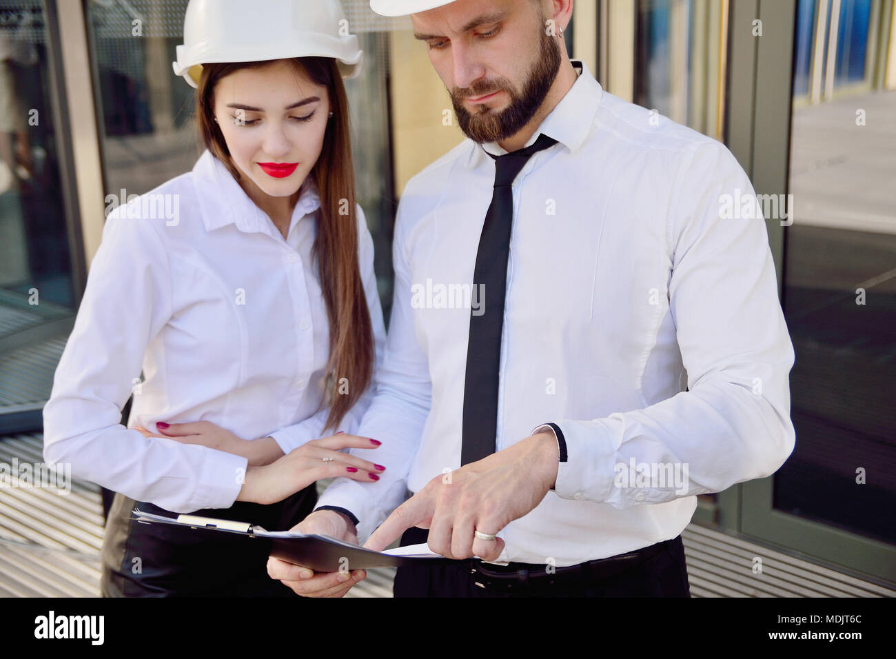 Una chica con un hombre en la construcción de cascos blancos y camisas  blancas con dibujos de manos contra un edificio de oficinas de cristal. Los  ingenieros. Planificación de la constr Fotografía