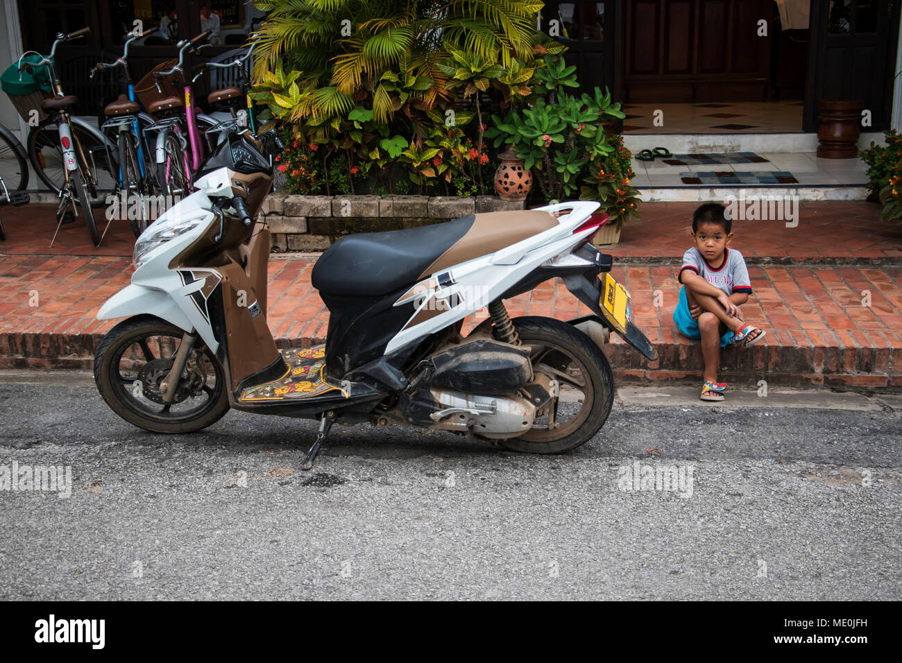 A lo largo de Calle Sisavangvong muchacho sentado junto a una motocicleta estacionada, Luang Prabang, Luang Prabang, Laos Foto de stock