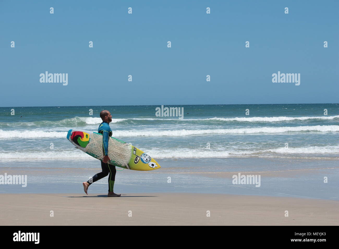 Surfista masculino llevando surfboard, vestida con traje de cuerpo completo caminando por la playa de Praia do Rosa, Brasil. Foto de stock