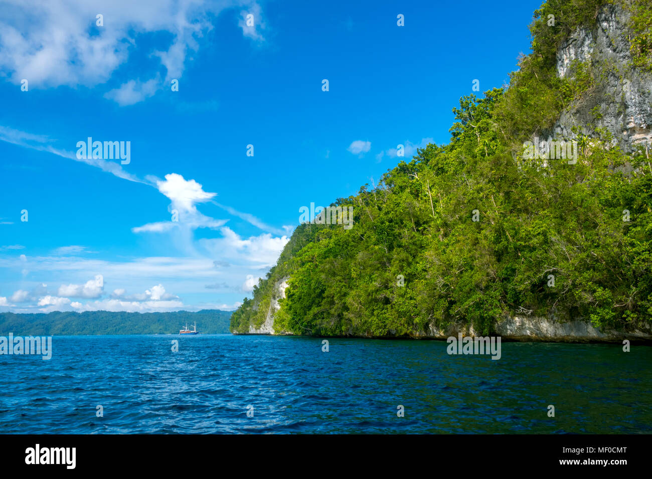 Indonesia. Costa rocosa de una isla tropical en clima soleado. Selva tropical en una pendiente. Velero en la distancia Foto de stock