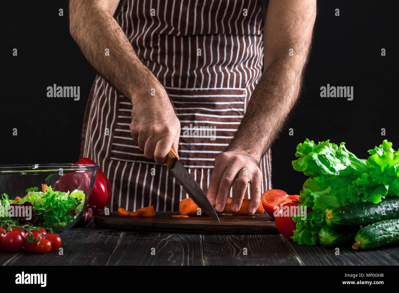 Casa joven hombre Cook en el delantal trocear la zanahoria con cuchillo de cocina. Foto de stock
