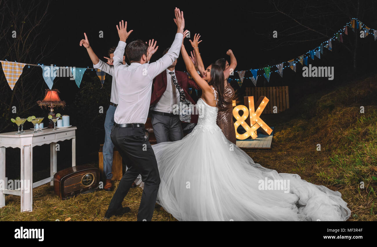 Feliz novia y amigos con los brazos levantados bailando y divirtiéndose en una noche de fiesta de campo Foto de stock