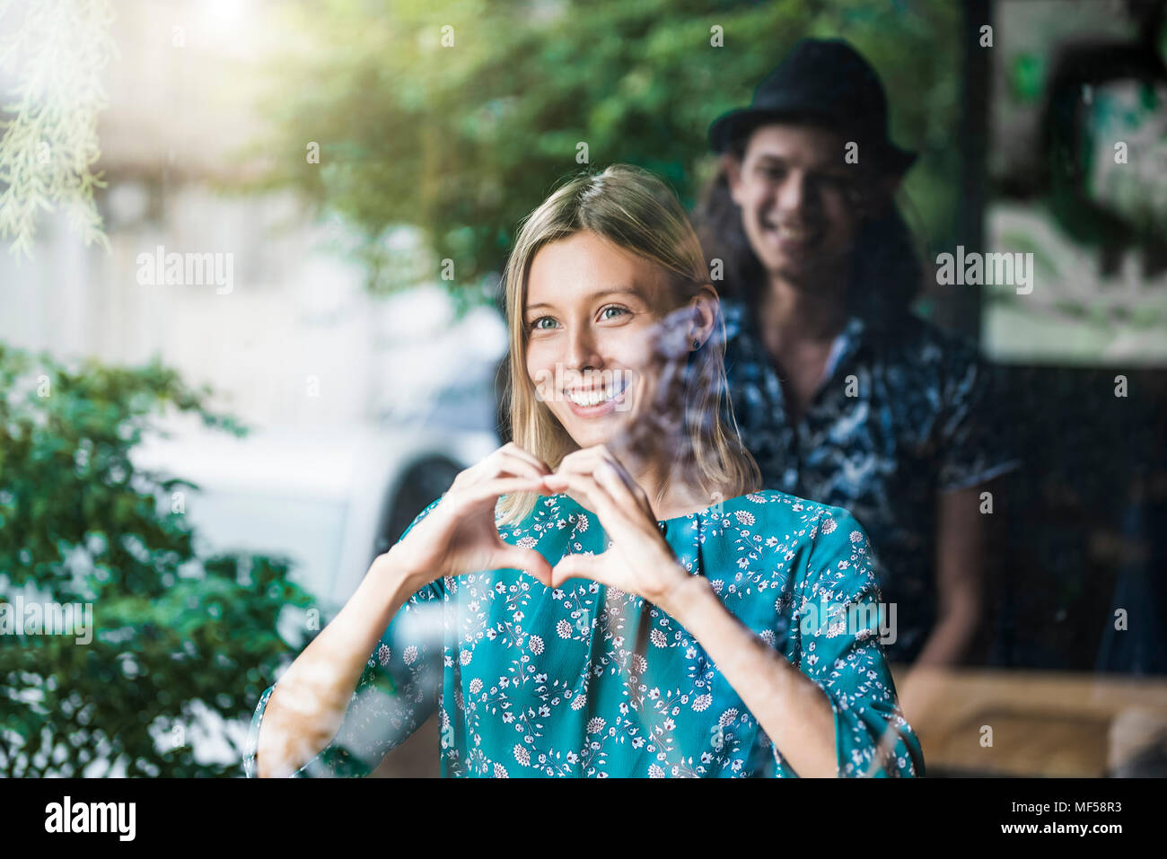 Hermosa joven haciendo un dedo corazón del bastidor y sonriente a su novio a través de una ventana Foto de stock