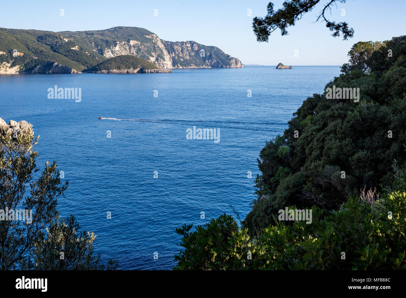 Vistas a un paisaje de mar bahía desde una ubicación elevada en Corfú, Grecia, con aguas azules, verdes montañas, pequeña isla y un barco rápido con dos personas en ella Foto de stock