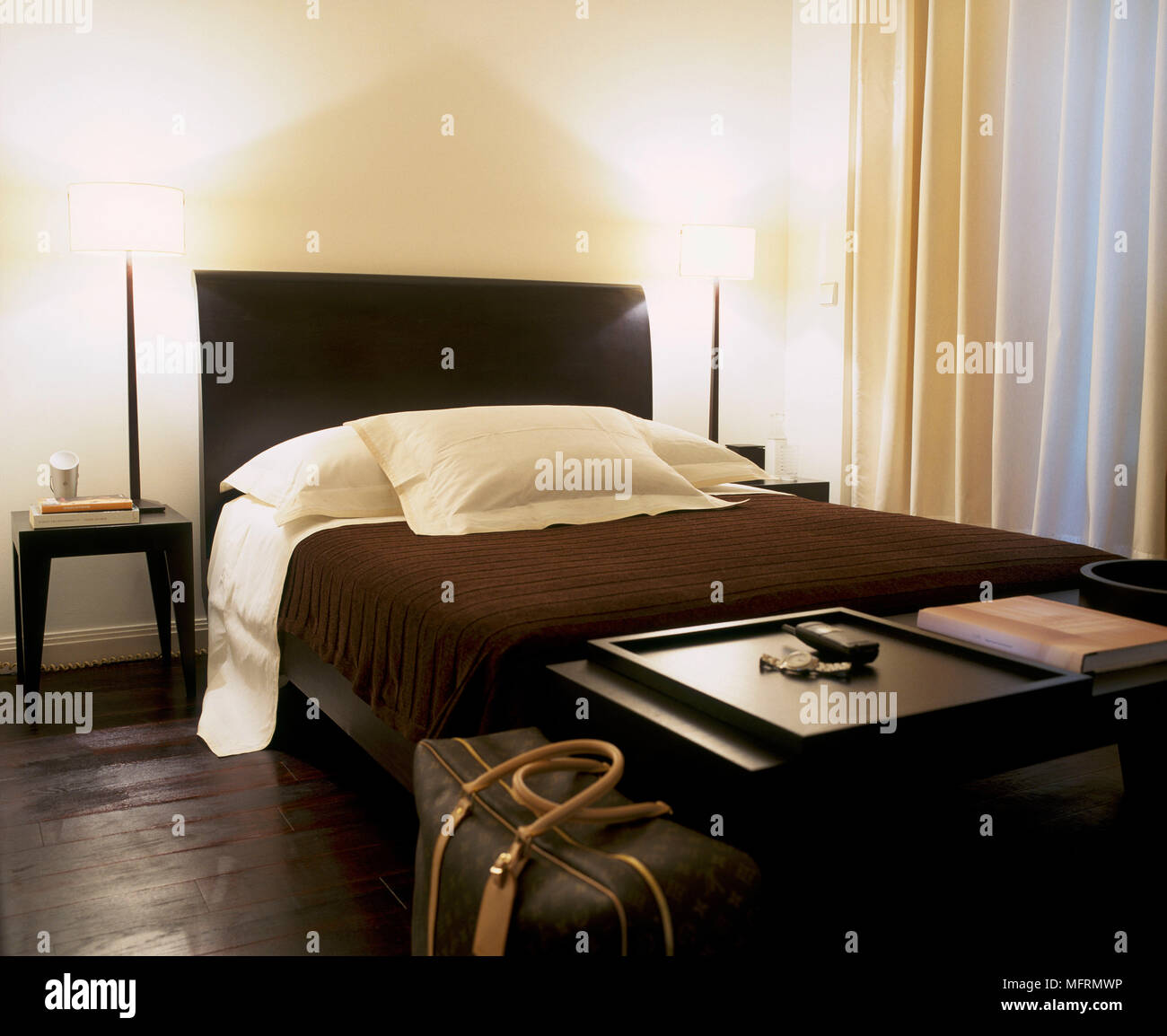 Un moderno dormitorio con una cama doble con cabecero tapizado brown bedcover mesa lateral piso de madera par de luces encendidas Foto de stock