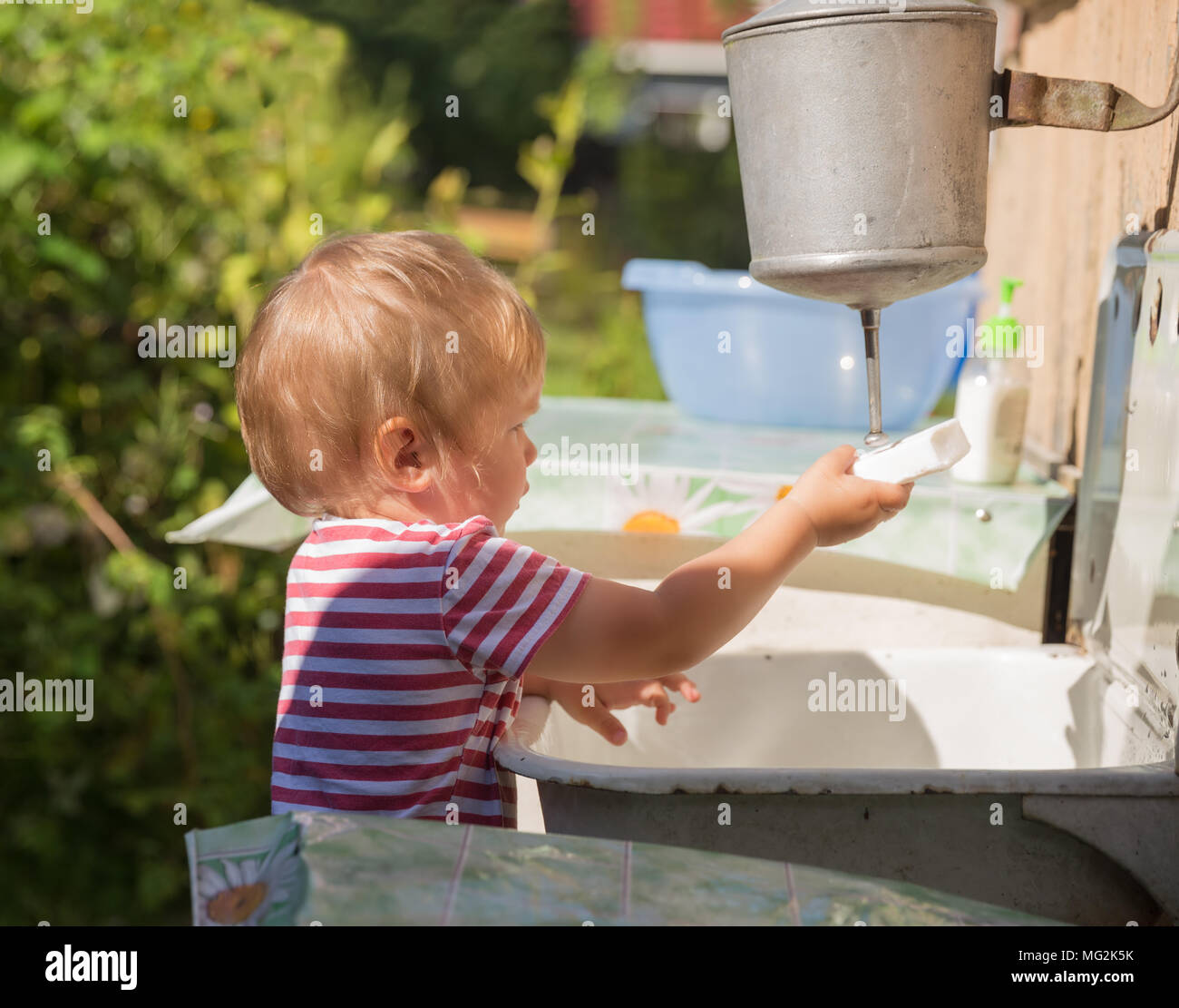Un niño pequeño, pequeño, de 2 años, rubio, de camiseta de rayas se lava las manos bajo lavabo afuera en el campo Foto de stock