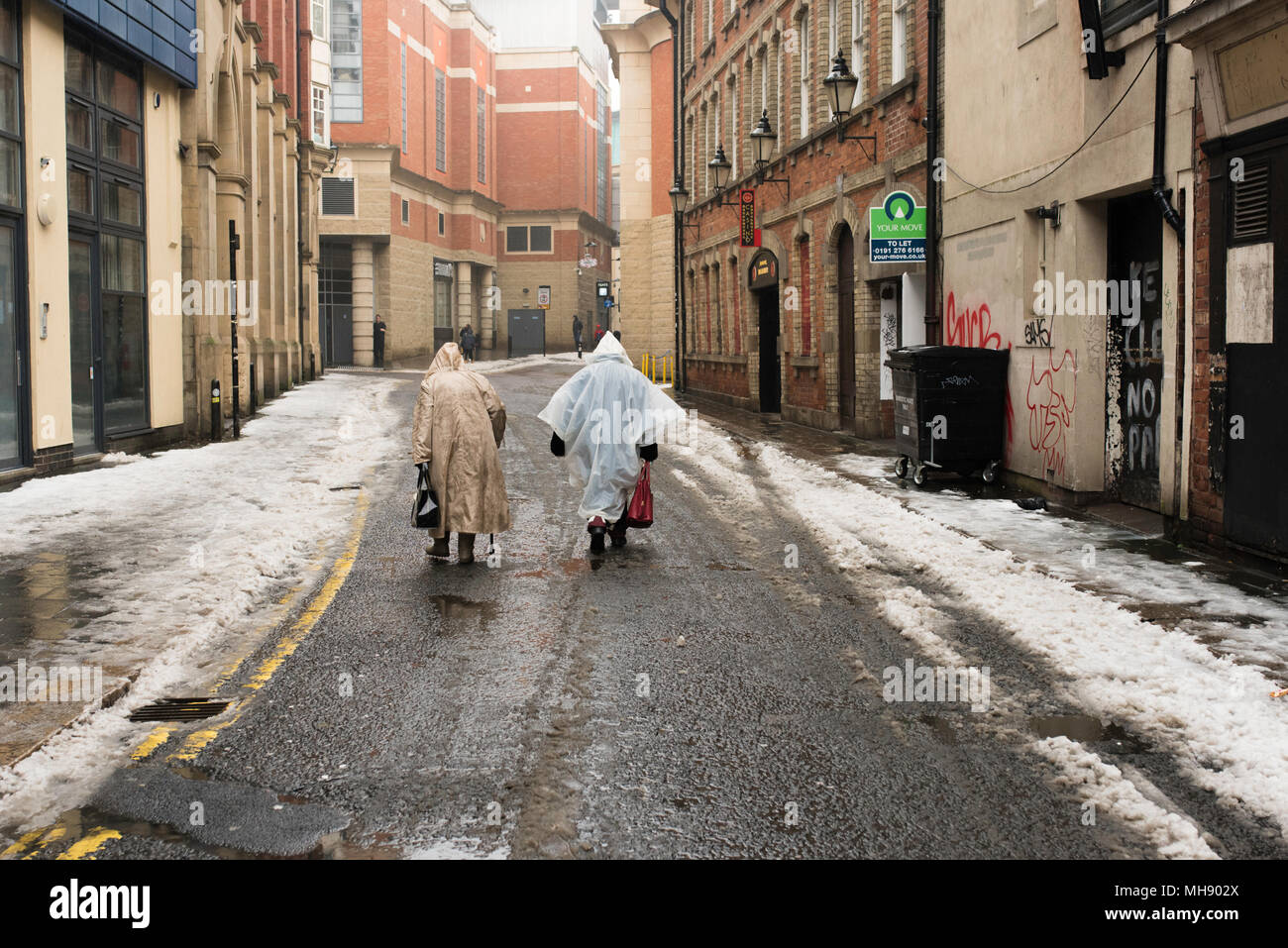 Dos ancianas vistiendo impermeables de plástico caminando por las calles nevadas de la ciudad de Newcastle, Tyne y desgaste, Reino Unido. Foto de stock
