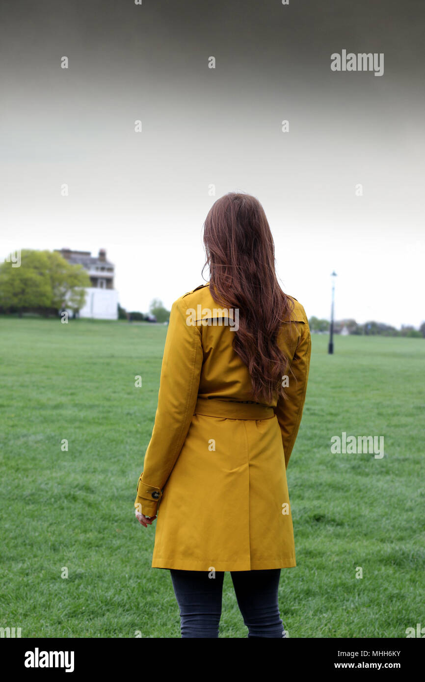 Mujer joven de pie en un parque con su espalda a la cámara mirando al horizonte, ella lleva un impermeable color ocre. Foto de stock