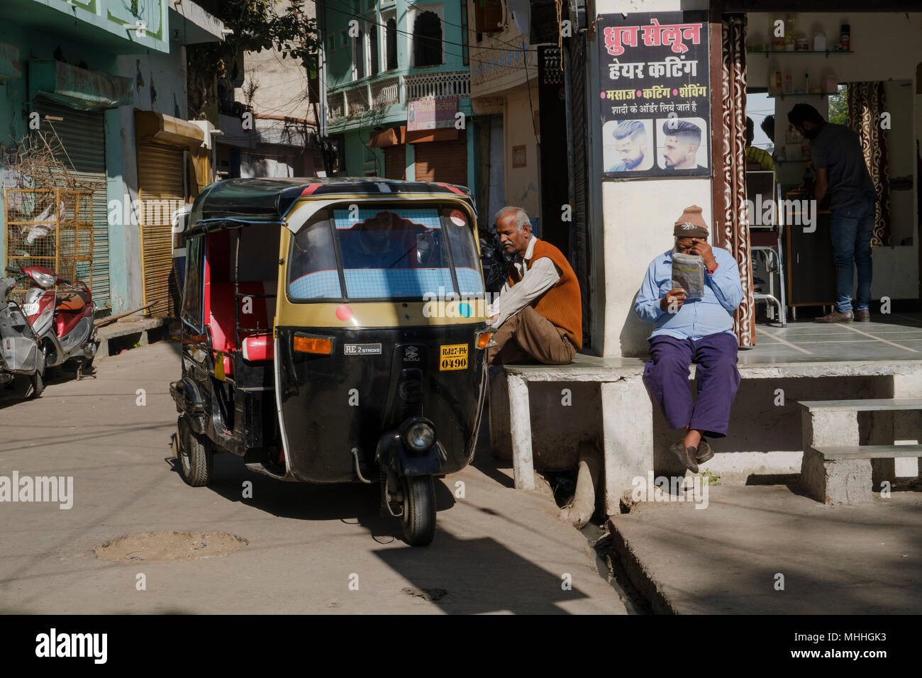 Hombres sentados fuera de peluquería junto a un tuk tuk y leyendo un periódico. Udaipur, también conocida como la ciudad de Lagos, la Venecia de Oriente, es la capital histórica del reino de Mewar, Rajasthan. Foto de stock