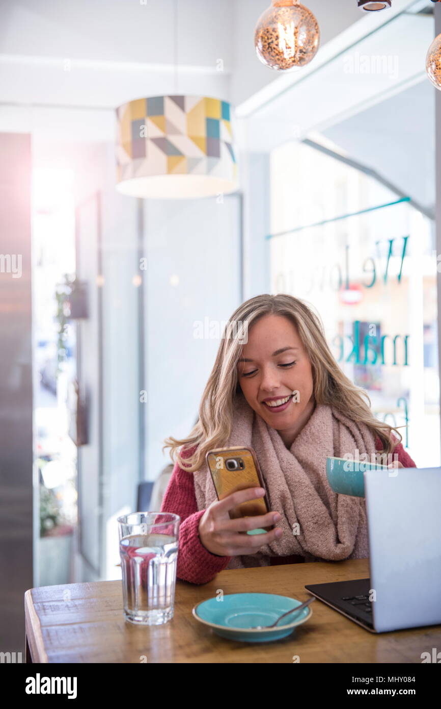 Mujer sentada en la cafetería, mirando a smartphone, sosteniendo la taza de café Foto de stock