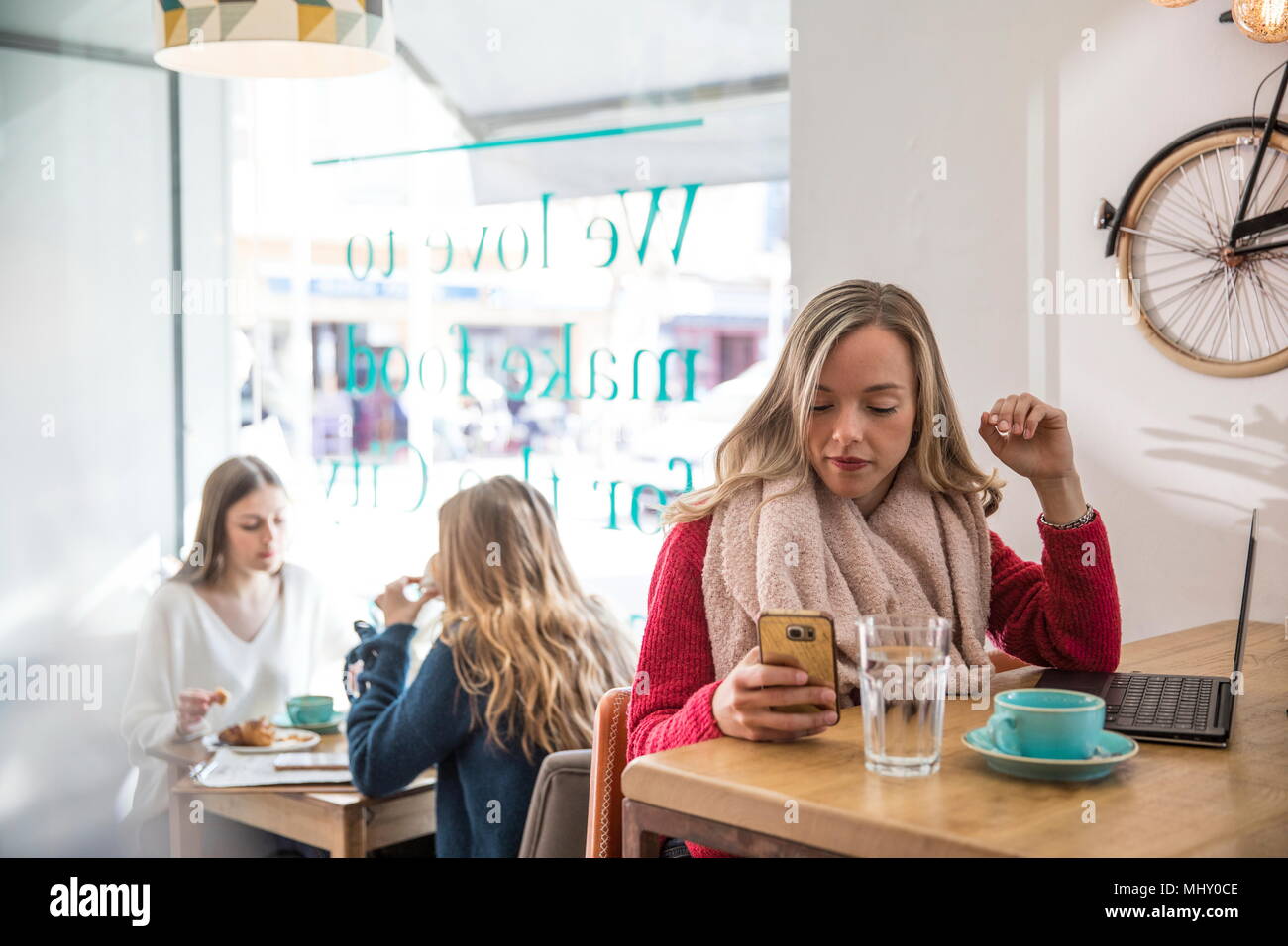 Mujer sentada en la cafetería, mirando a smartphone, laptop en frente de ella Foto de stock