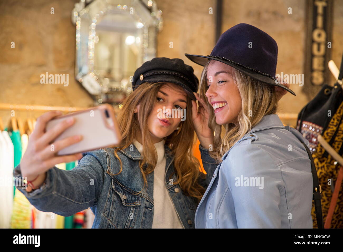 Dos amigos en la tienda, probando sombreros, teniendo selfie, utilizando el smartphone Foto de stock