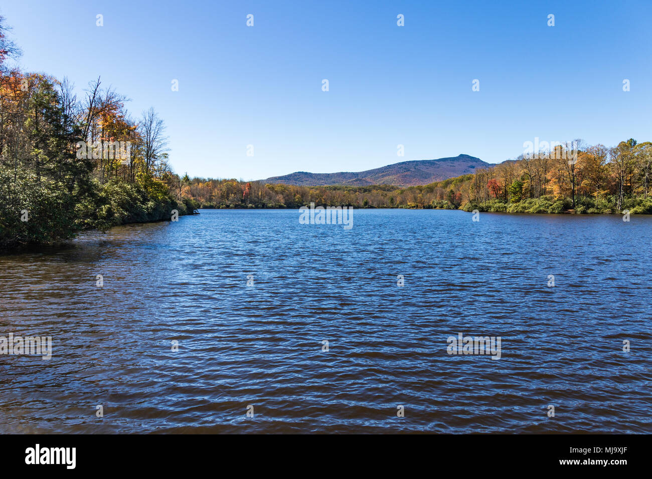 En el otoño de un tranquilo lago de montaña rodeado de color en el otoño y el cielo azul, y una línea de cresta de la montaña en la distancia. Foto de stock