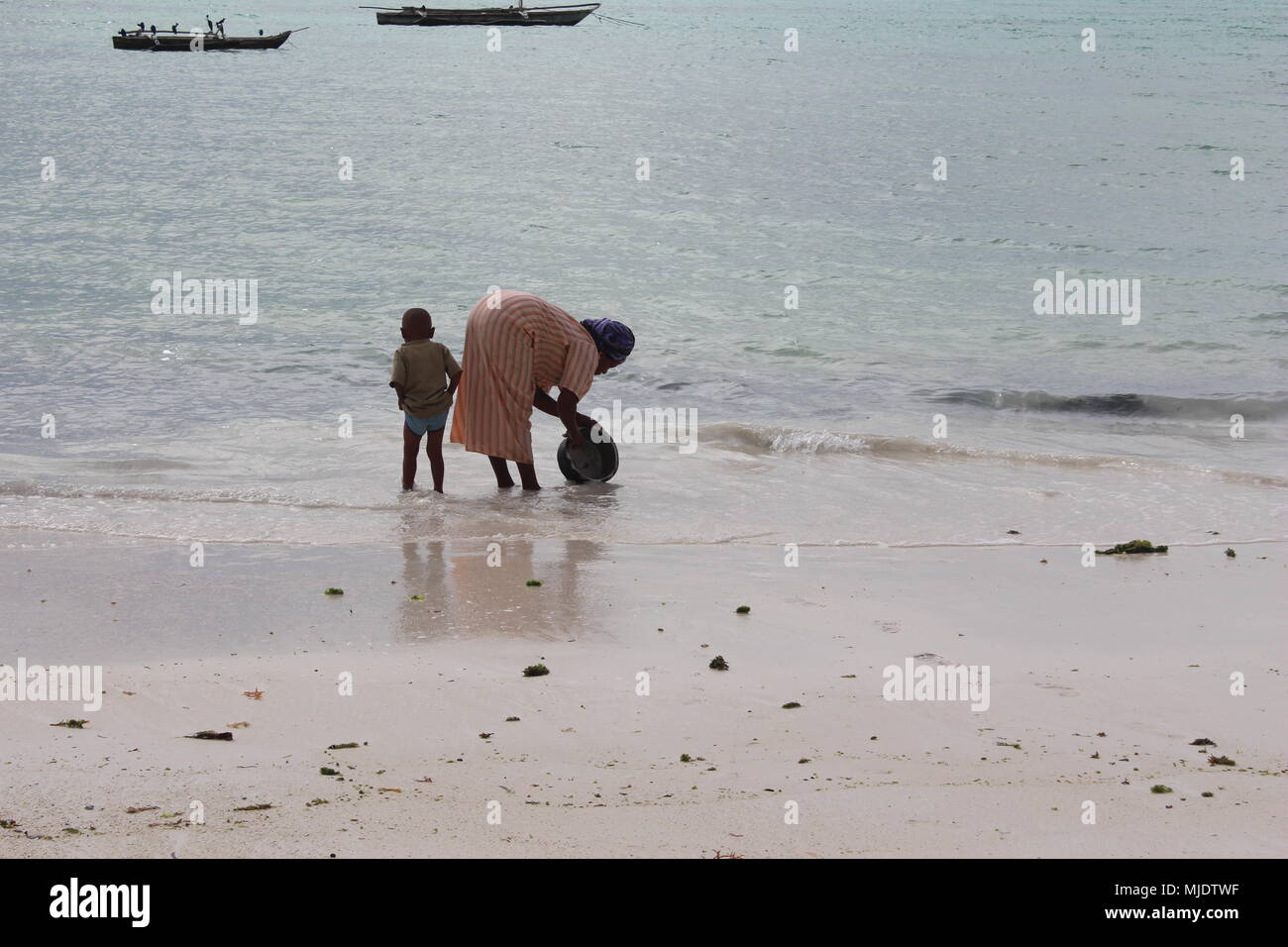 Mujer Africana lavando ollas con arena en la playa, mientras que su joven hijo tira sus cortos hacia arriba para que no se moje. Zanzíbar, Tanzania. Foto de stock