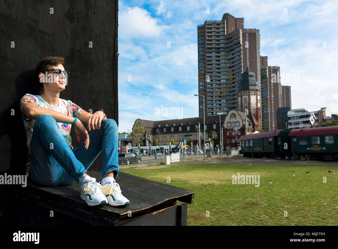 Joven con gafas de sol, sentado, apoyado contra la pared, ciudad de rascacielos, Foto de stock