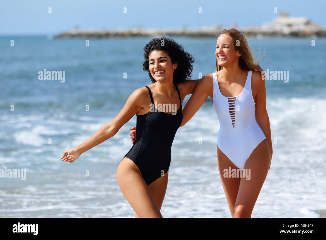 Dos mujeres jóvenes con bellos cuerpos en de baño en una playa tropical. Gracioso y caucásica árabe mujeres vistiendo trajes baño blanco y negro caminando al Fotografía de stock -