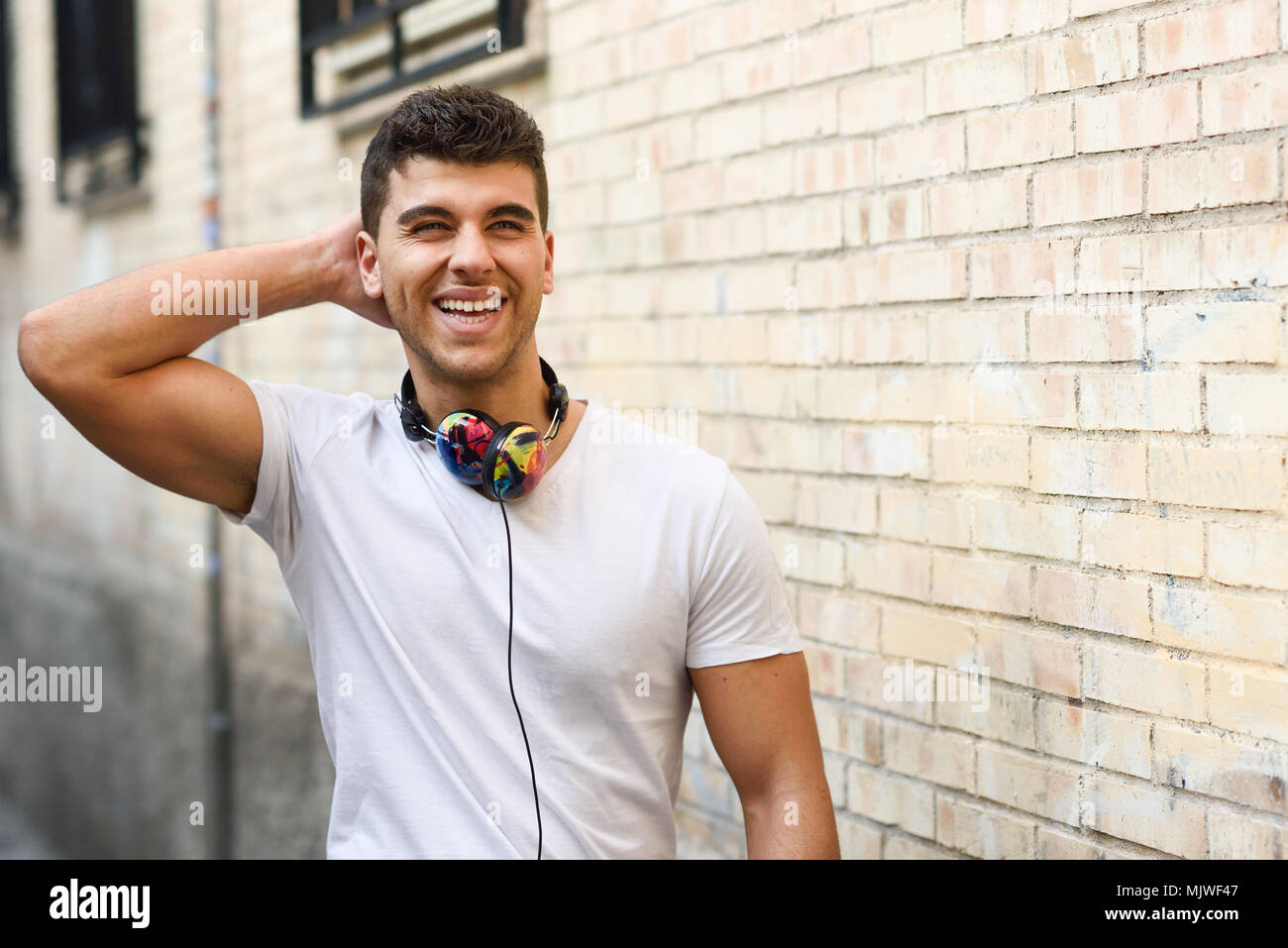 Retrato de joven en el contexto urbano de escuchar música con auriculares. Vestían camiseta blanca cerca de una pared de ladrillo Foto de stock