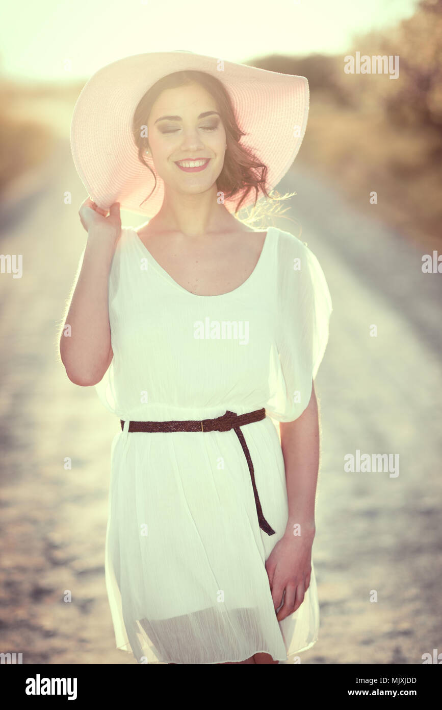 Retrato de mujer joven con los ojos cerrados en una carretera rural en la  primavera. Vestida de vestido blanco, cinturón marrón y rosa sombrero para  el sol. Retroiluminación photogra Fotografía de stock -